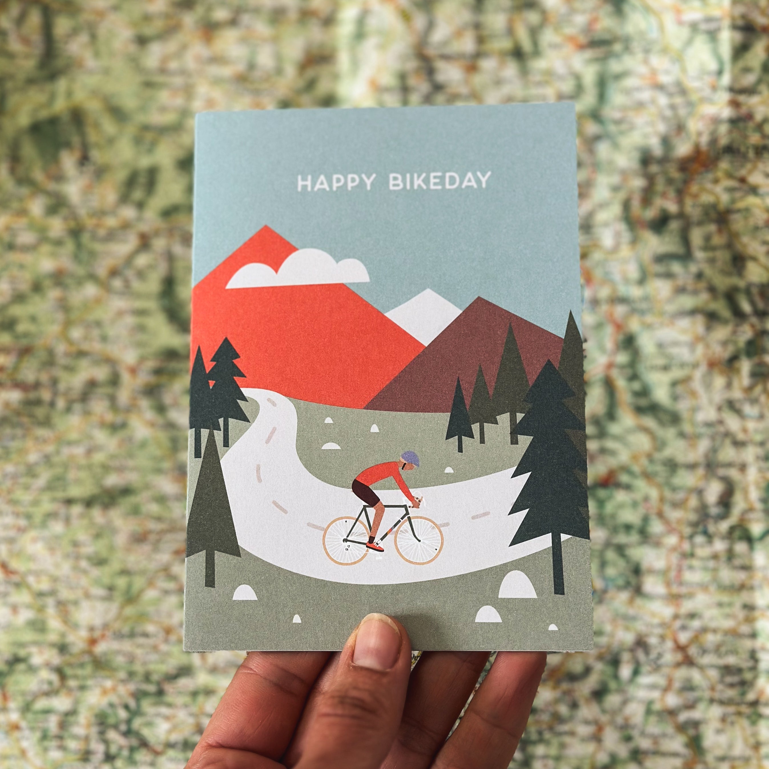 Grußkarte von Roadtyping mit der Illustration eines Radfahrers, der eine Bergstraße hochfährt. Darüber steht der Text "Happy Bikeday".