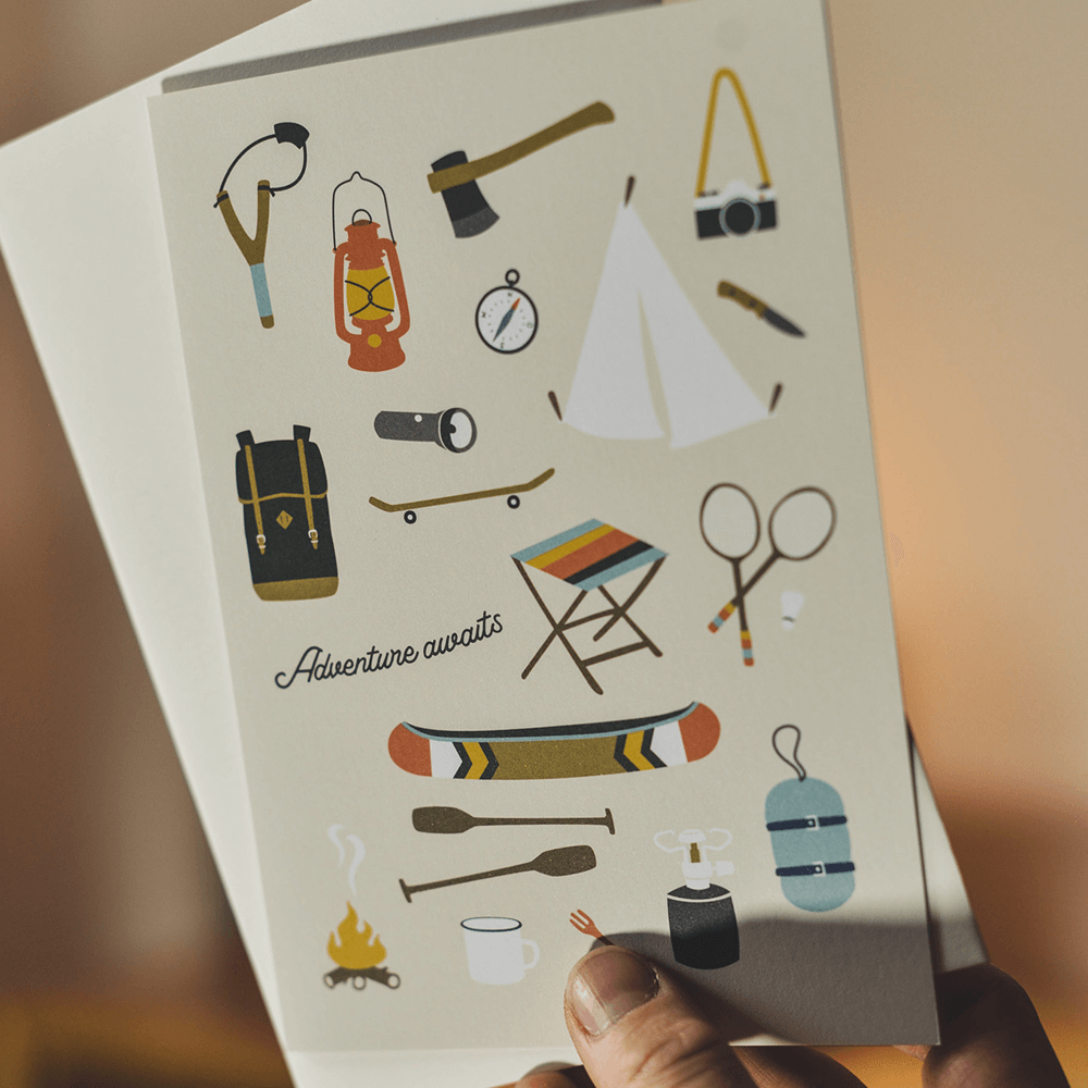 Grußkarte von Roadtyping mit der Illustration eines Kanus, umgeben von Outdoor-Utensilien wie einer Axt, einem Lagerfeuer und einem Becher.