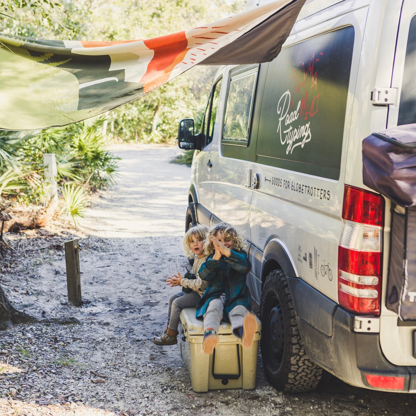 Zwei Kinder sitzen unter dem Sonnensegel vor dem Wohnmobil